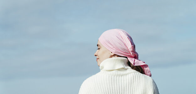 Close up photo of back view of young woman with cancer. She is wearing a pink scarf on her head and white jersey. She is feeling the sun on her face. She is looking to the left. Blue sky on background.